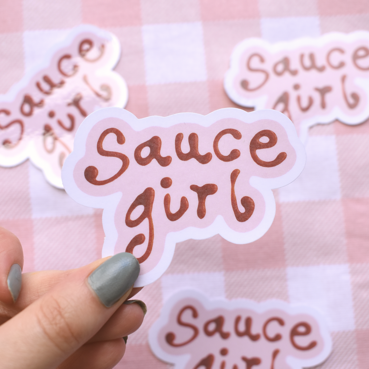 Sticker with 'Sauce girl' text held by a hand against a pink checkered background