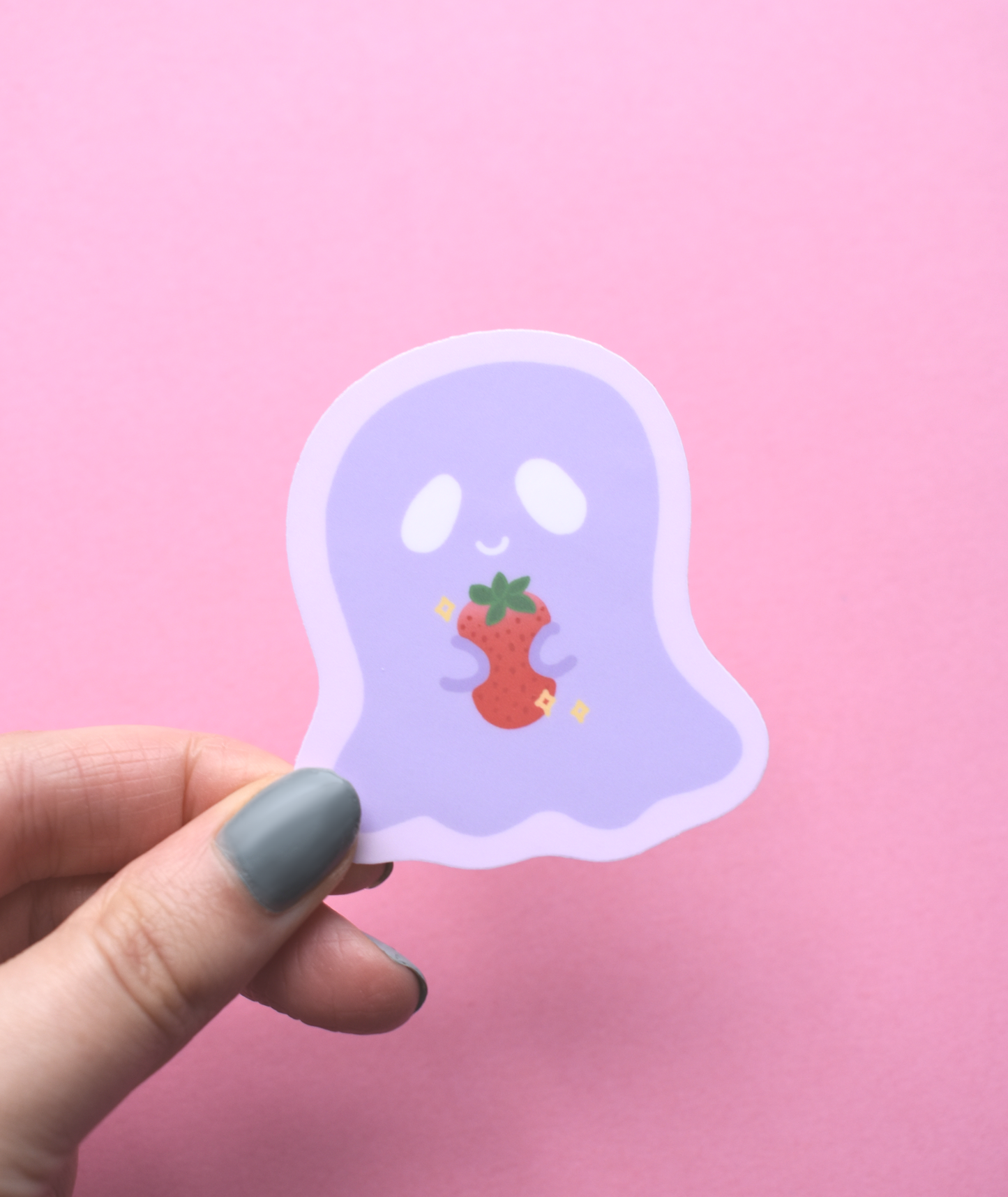 Hand holding a ghost-shaped sticker with a strawberry on a pink background