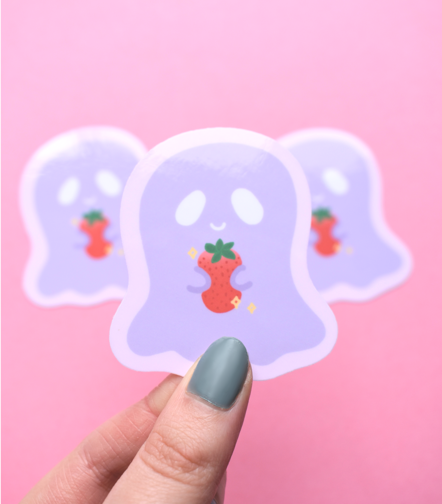 Three ghost-shaped stickers with strawberry designs on a pink background, held by a hand with green nail polish.