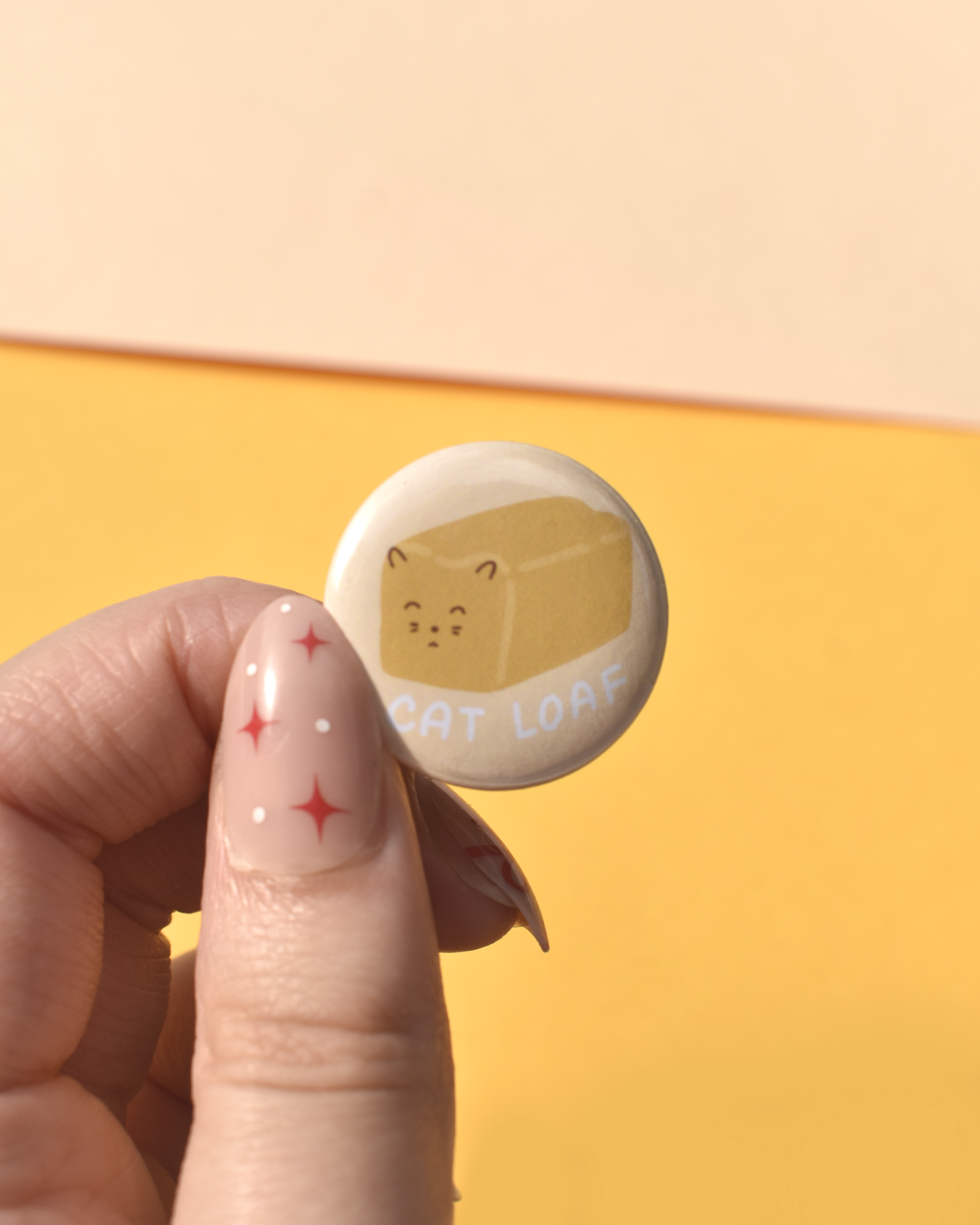 Button badge with cat loaf design held by a hand against a yellow background