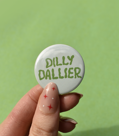 Button badge with 'Dilly Dallier' text held by a hand against a green background