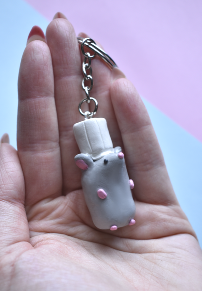 Hand holding a keychain with a small rat with a chef hat figure on a pink and blue gradient background