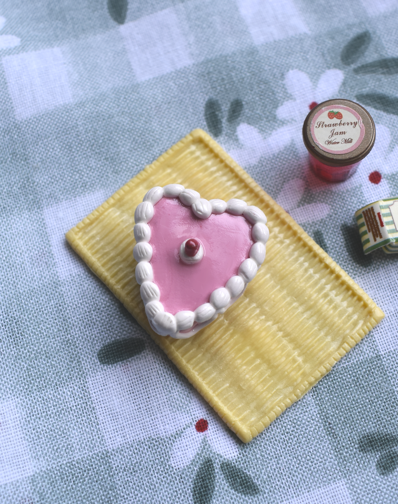 Heart-shaped pink cake magnet with white frosting on a yellow square base, placed on a floral-patterned fabric background.