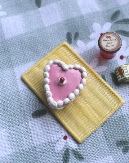 Heart-shaped pink cake magnet with white frosting on a yellow square base, placed on a floral-patterned fabric background.