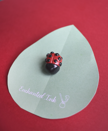 Lapel pin shaped like a ladybug on a green leaf with 'Enchanted Ink' branding on a red background