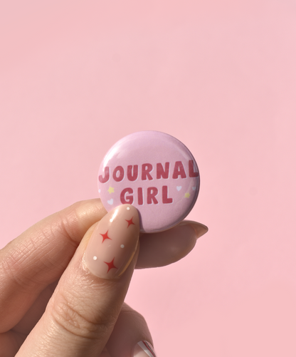 Pink button badge with 'Journal Girl' text held by a hand against a pink background
