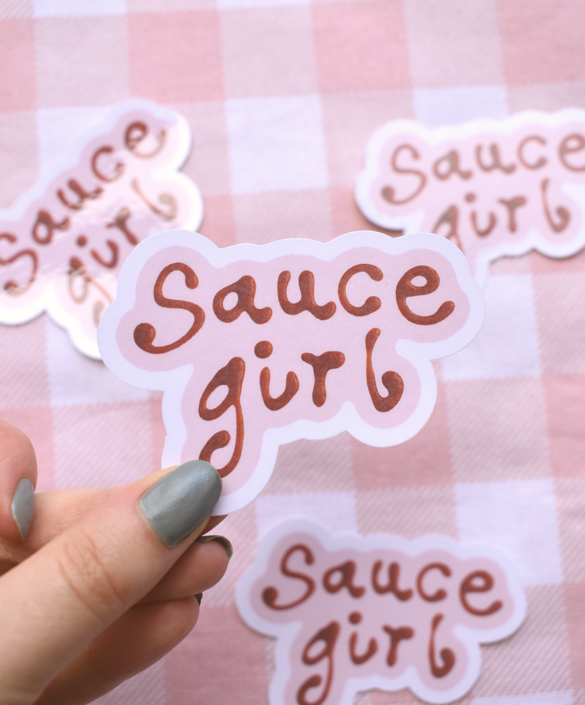 Sticker with 'Sauce girl' text held by a hand against a pink checkered background
