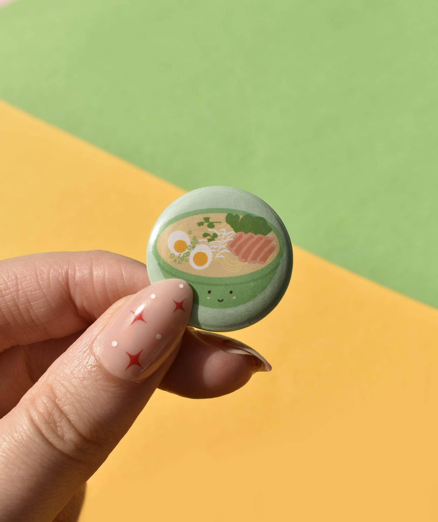 Hand holding a small round badge with a design of a bowl of ramen on a colorful background