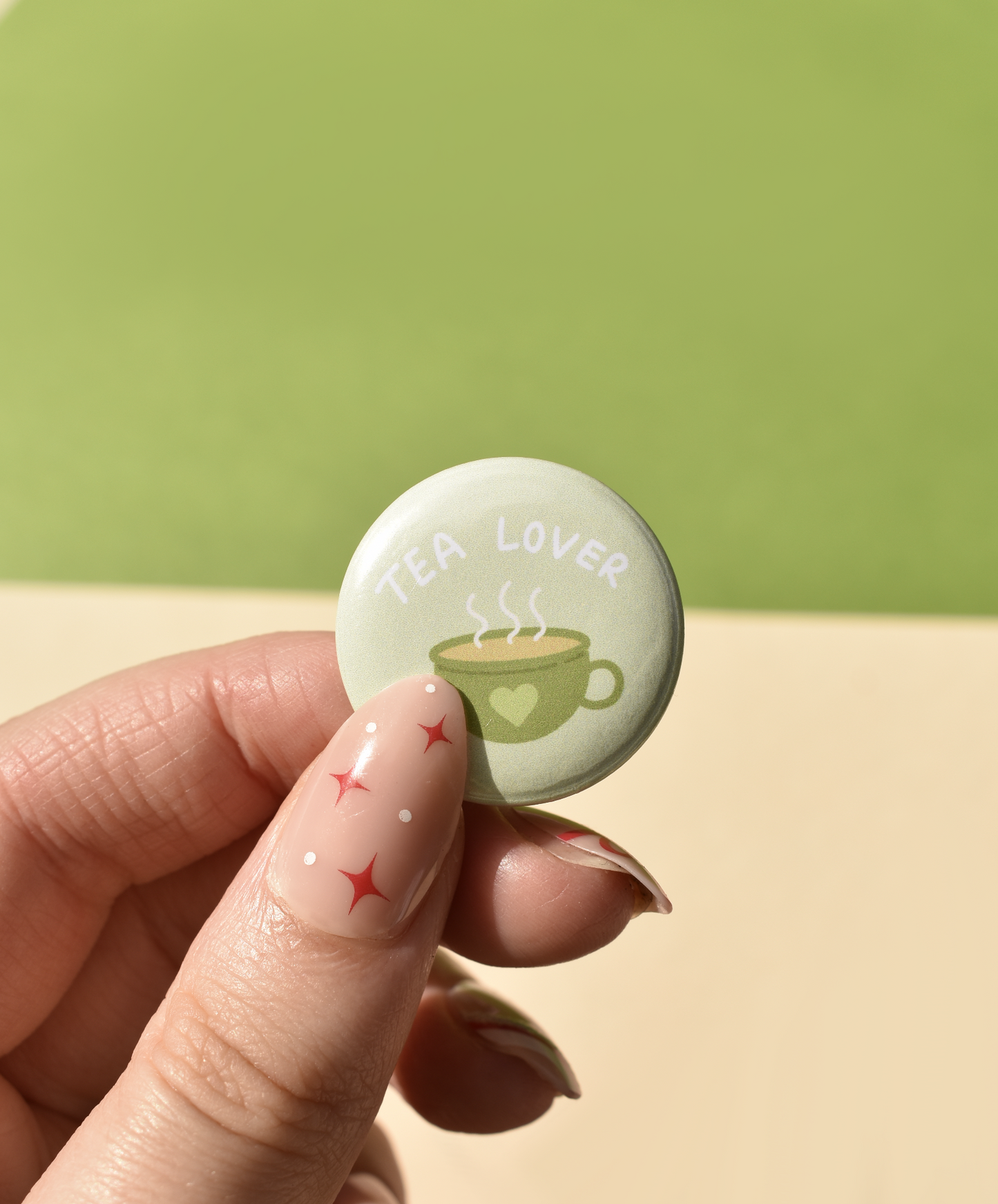 Button badge with 'Tea Lover' text and tea cup design held by a hand against a green background