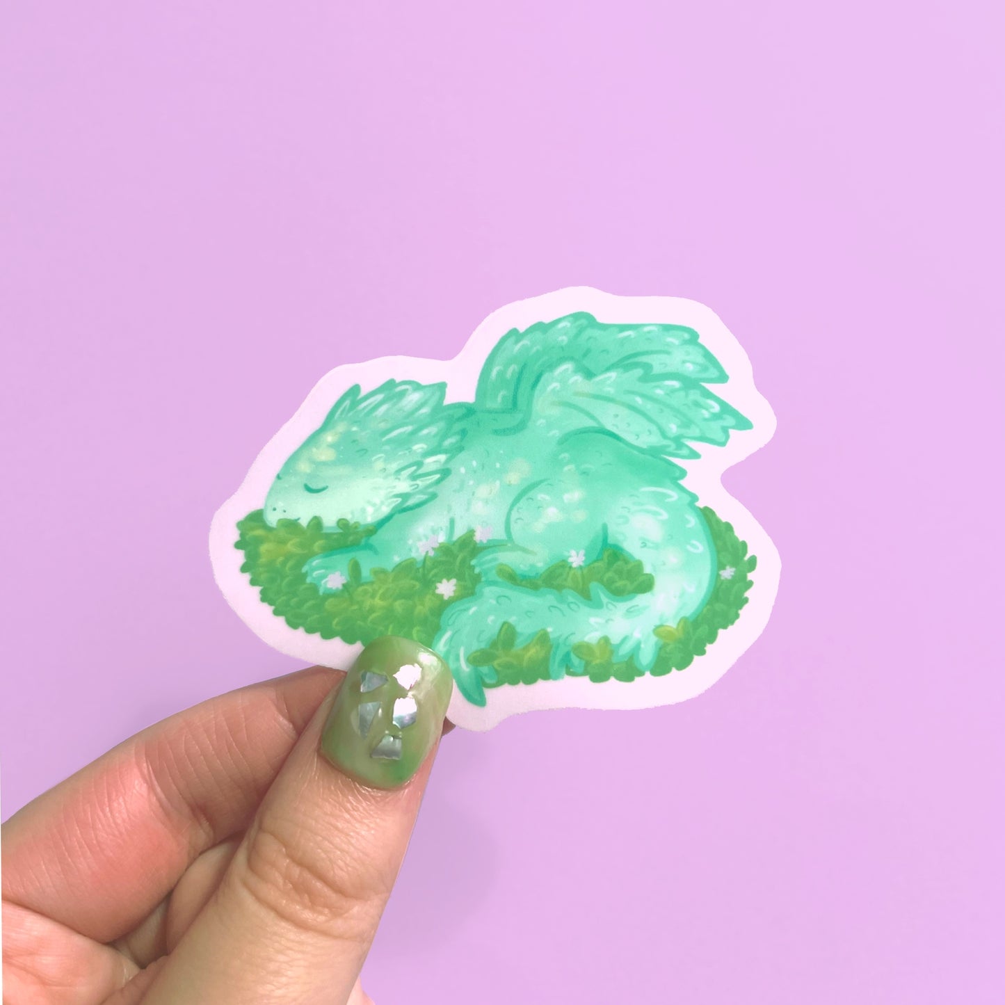 Cute fantasy dragon sticker featuring a baby green dragon sleeping peacefully on leafy bed