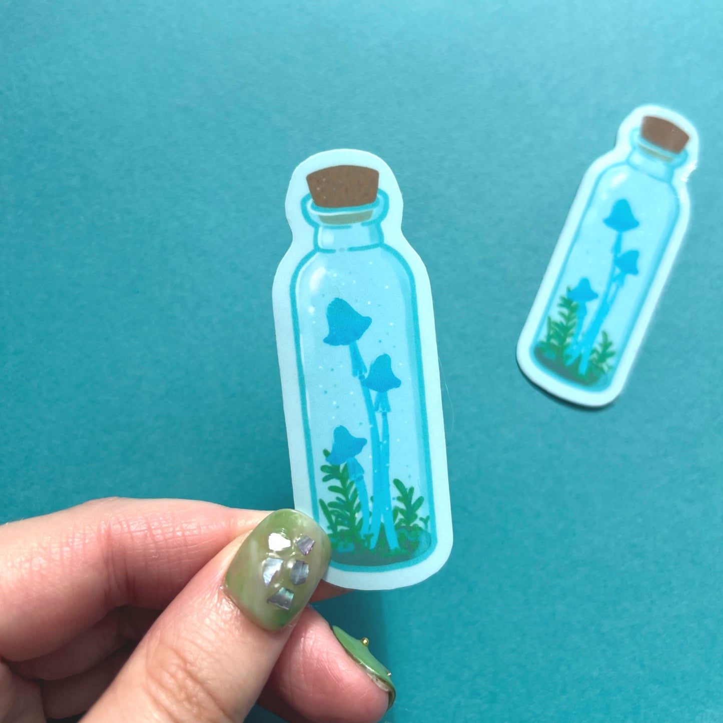 Whimsical fantasy sticker featuring blue mushrooms inside a clear potion bottle with greenery