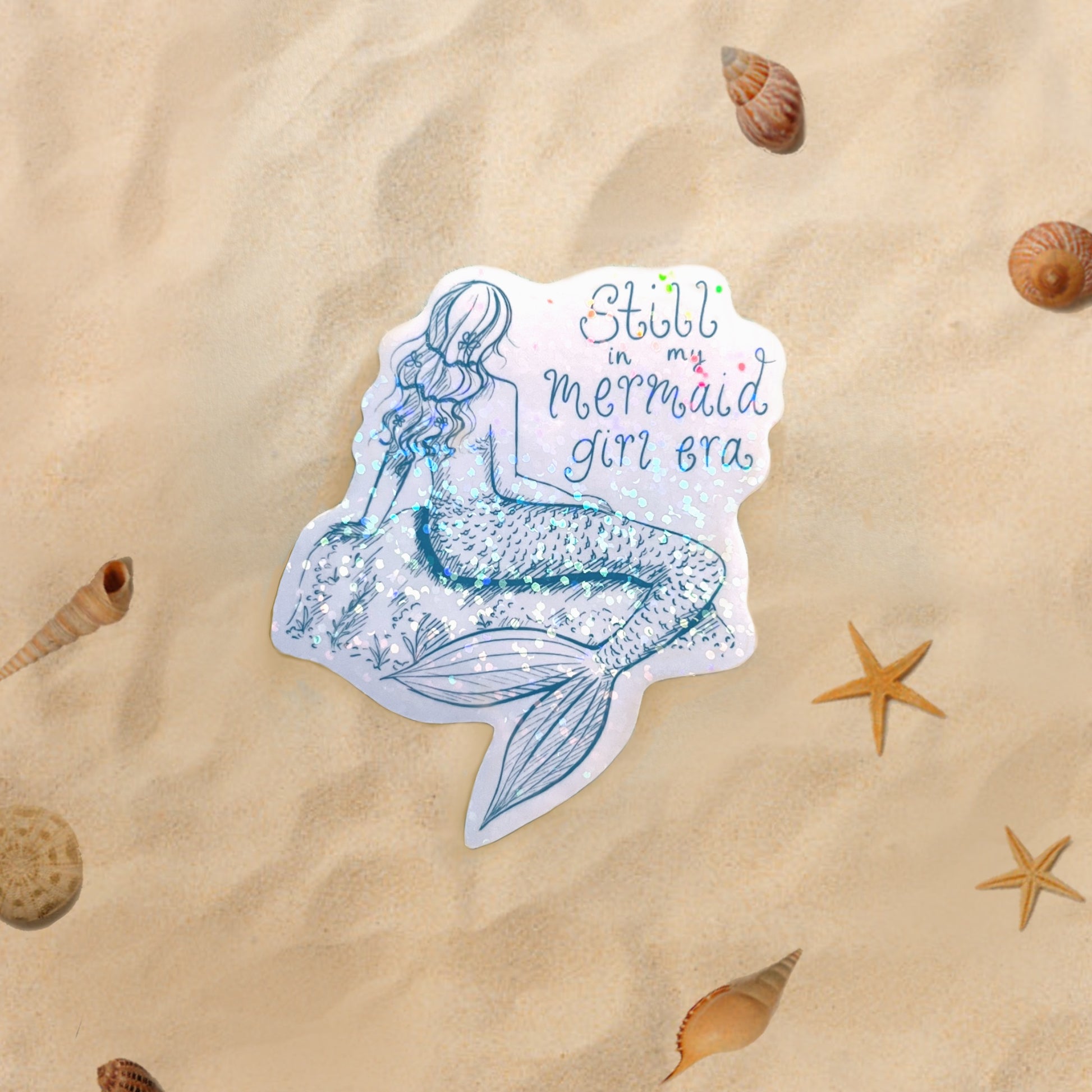 Mermaid sticker with 'Still in my mermaid girl era' text on sand with shells and starfish